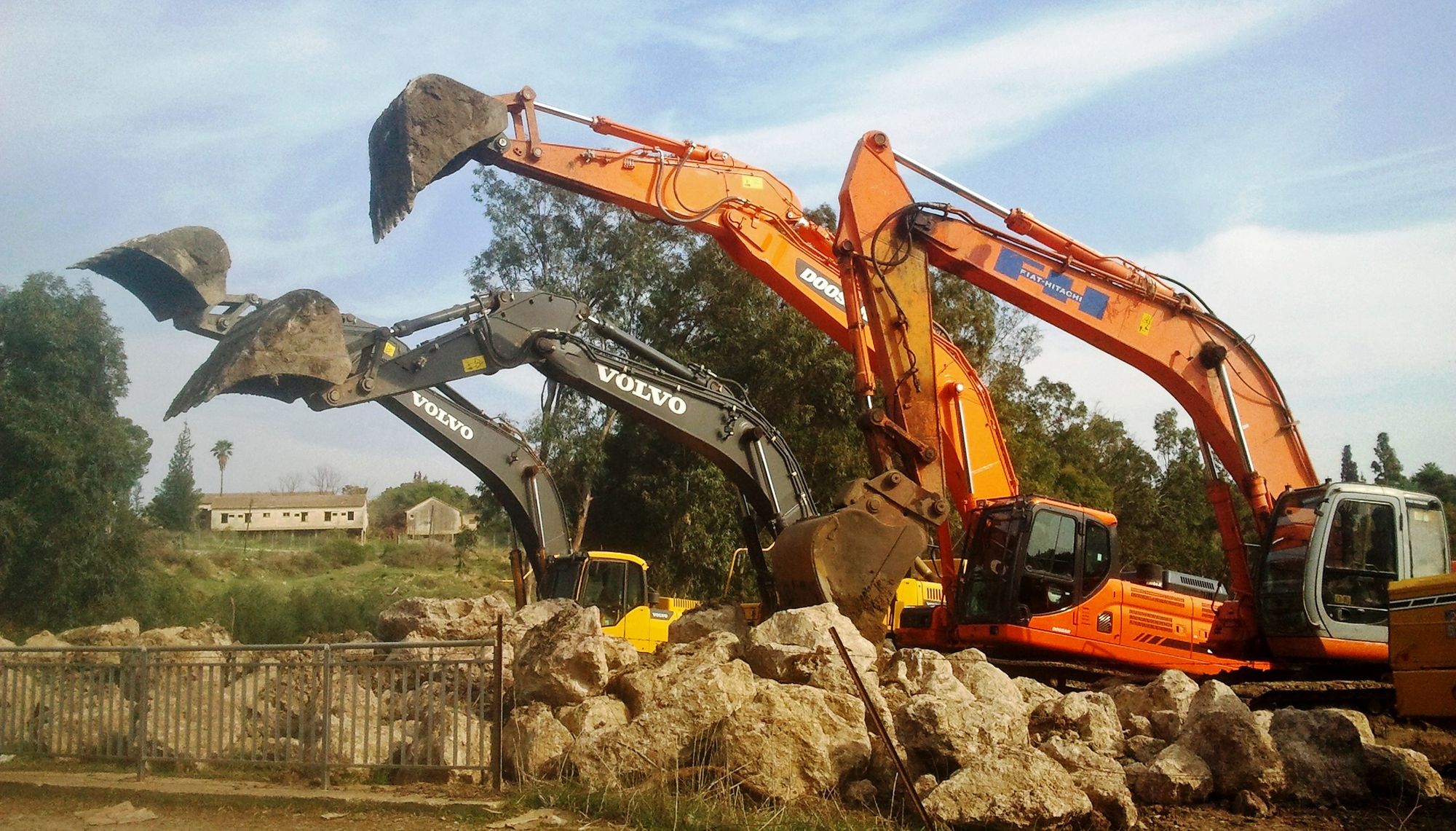 Four excavators parked side by side on a rocky site, buckets raised
